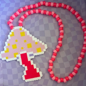 Puede incluir: Un collar de cuentas rosas y rojas con un colgante de hongo. El colgante de hongo es rosa claro con cuadrados amarillos y un tallo rojo. El collar está hecho de cuentas rosas y rojas.