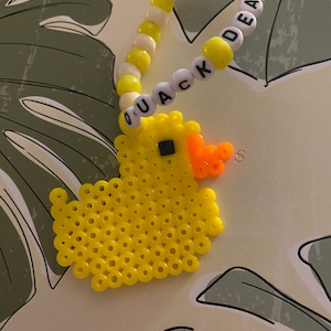 May include: A yellow rubber ducky made of perler beads with a black eye and an orange beak. The ducky is next to a white and yellow beaded bracelet with the words "QUACK DEALER" spelled out.