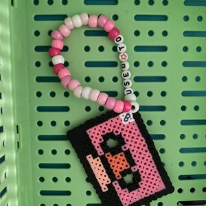 Puede incluir: Un brazalete de cuentas rosa y blanco con la palabra "USED TO" escrita en él. El brazalete está al lado de un diseño de arte de píxeles rosa y negro de una cinta de casete.