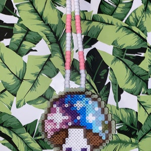 May include: A colorful beaded necklace with a pixelated mushroom charm. The mushroom is blue, purple, pink, and white with a brown stem and a white face. The necklace is made of pink and white beads.