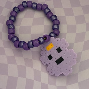 May include: A purple beaded bracelet with a charm. The bracelet is made of small, round, dark purple beads. The charm is a light purple cloud shape with black and yellow details. The background is a checkered pattern of light and dark purple.