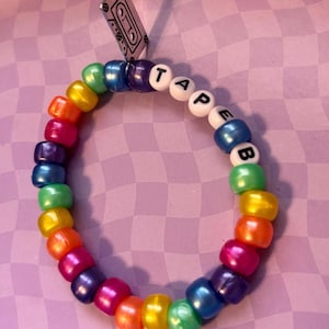 Pode incluir: Uma pulseira de contas coloridas com contas de arco-íris. A pulseira soletra a palavra "TAPE" com contas de letras brancas. Um pingente de fita cassete prateada pende da pulseira. Em um fundo roxo quadriculado.