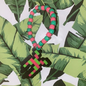 Puede incluir: Un brazalete de cuentas rosa y verde con un dije pixelado negro y rosa. El dije tiene forma de letra H.