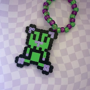 Puede incluir: Un dije de figura pixelada verde y negra con una pulsera de cuentas verdes y moradas a juego. El dije y la pulsera están sobre un fondo a cuadros morado y blanco. El dije tiene un diseño pixelado.