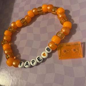 Pode incluir: Pulseira de contas laranja com a palavra "TAPE B" escrita em contas de letras brancas. A pulseira inclui um berloque em forma de fita cassete. As contas são uma mistura de laranja sólido e laranja transparente com flocos dourados.