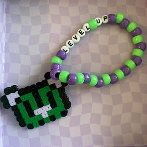 Puede incluir: Una pulsera de cuentas con las palabras "LEVEL UP" escritas con cuentas de letras blancas. La pulsera presenta cuentas verdes y moradas. Un dije pixelado verde y negro está unido a la pulsera.
