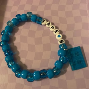Pode incluir: Uma pulseira de contas azuis com a palavra "TAPE" escrita em contas de letras brancas. Um pequeno pingente de fita cassete azul pende da pulseira. A pulseira está em uma superfície xadrez roxa e branca.