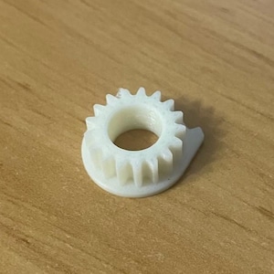 Puede incluir: Un pequeño engranaje de plástico blanco con un borde exterior dentado circular y un agujero central. El engranaje tiene una pequeña extensión curva en un lado. El objeto está sobre una superficie de madera.