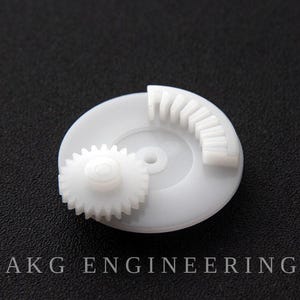 Puede incluir: Un conjunto de engranajes de plástico blanco que presenta una base circular con una pista de engranaje parcial elevada y un engranaje circular más pequeño y separado colocado encima. El texto AKG ENGINEERING aparece en blanco al pie de la imagen.