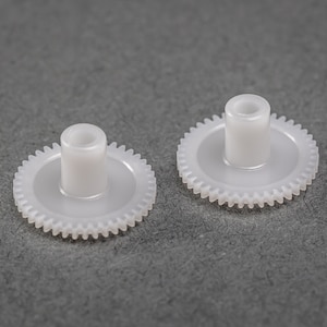 Könnte beinhalten: Zwei weiße Kunststoffzahnräder mit einem zentralen Loch. Die Zahnräder haben eine glatte, abgerundete Oberfläche und eine Reihe von Zähnen am Außenrand.
