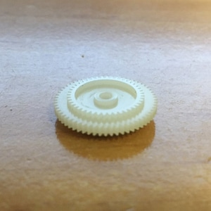 Può includere: Un ingranaggio in plastica bianca con un foro centrale. L'ingranaggio ha una serie di denti attorno al bordo esterno.