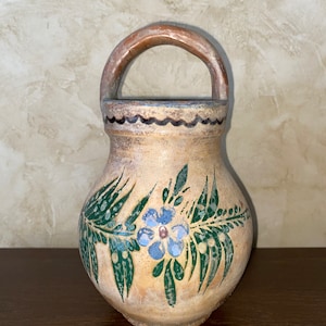 Può includere: Un vaso in ceramica dipinto a mano con manico. Il vaso è beige con un motivo floreale blu e verde. Il manico è marrone. Il vaso ha un bordo nero ondulato vicino alla parte superiore.