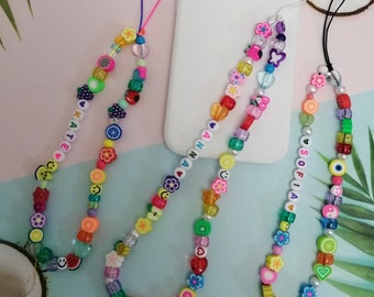 May include: Brightly coloured beaded phone charms with fruit, flower, and heart-shaped beads. Each charm has a name spelled out with white letter beads. The charms are in various colours, including pink, green, yellow, and orange.
