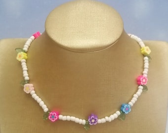 Op de afbeelding: Een witte kralenketting met kleurrijke bloemenbedels. De ketting heeft roze, gele, blauwe en paarse bloemen met groene bladeren.