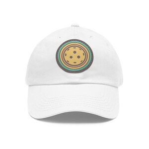 May include: White baseball cap with a colorful circular patch featuring a yellow pickleball with brown and blue accents.