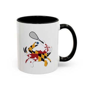 Könnte beinhalten: Weiße Keramik-Kaffeetasse mit schwarzem Innenraum und Henkel. Die Tasse zeigt eine Silhouette eines Krebses, der einen Tennisschläger und einen Ball hält, alles in den Farben der Flagge des Bundesstaates Maryland.