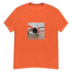 May include: Orange t-shirt featuring a square image of an airplane engine and part of the plane's body. The plane is white and orange. The engine is black and gray. A yellow traffic cone is in front of the engine.