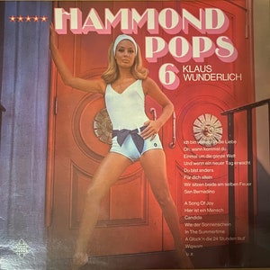 May include: A vintage vinyl record cover featuring a woman in a white swimsuit standing in front of a red door. The cover is titled "Hammond Pops 6" and features the artist's name, "Klaus Wunderlich". The cover also includes a list of songs, including "A Song of Joy", "Candida", and "Wigwam".