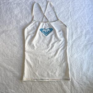 Könnte beinhalten: Ein weißes Tanktop mit dünnen, überkreuzten Trägern und einem hellblauen Roxy-Logo auf der Brustmitte. Das Kleidungsstück hat eine schlichte, körpernahe Passform mit dezenten, schillernden oder mehrfarbigen Nähten entlang der Kanten.