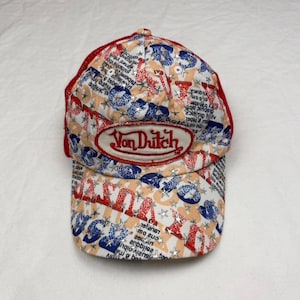 May include: A Von Dutch baseball cap with a red mesh back and a curved brim. The cap features a repeating pattern of blue, red, and white graphics, with the Von Dutch logo prominently displayed on the front.