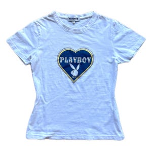 Peut inclure: T-shirt blanc avec un motif en forme de cœur bleu portant l'inscription "PLAYBOY" et un logo de lapin. Le t-shirt a des manches courtes et un col rond. L'étiquette de taille indique "M".
