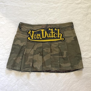 May include: Camouflage print pleated skirt with the text "Von Dutch" in yellow and black. The skirt is made of a durable fabric and features a classic pleated design.