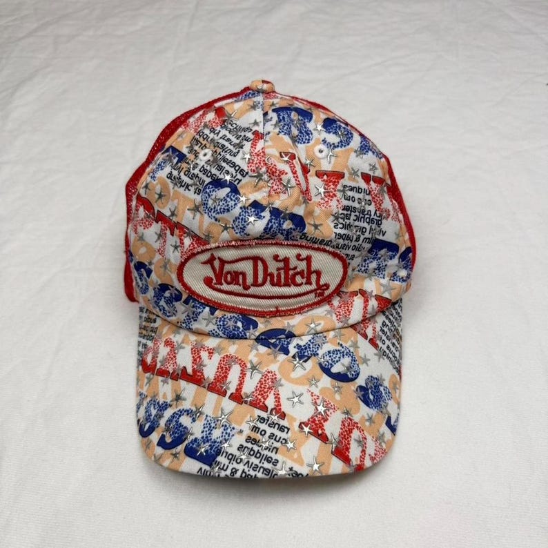 May include: A Von Dutch baseball cap with a red mesh back and a curved brim. The cap features a repeating pattern of blue, red, and white graphics, with the Von Dutch logo prominently displayed on the front.