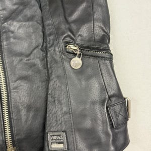 May include: Black leather jacket with a perforated design. Features a silver zipper, a small zippered pocket, and a decorative silver charm. The jacket has a rectangular label with the Versace Jeans Couture logo.