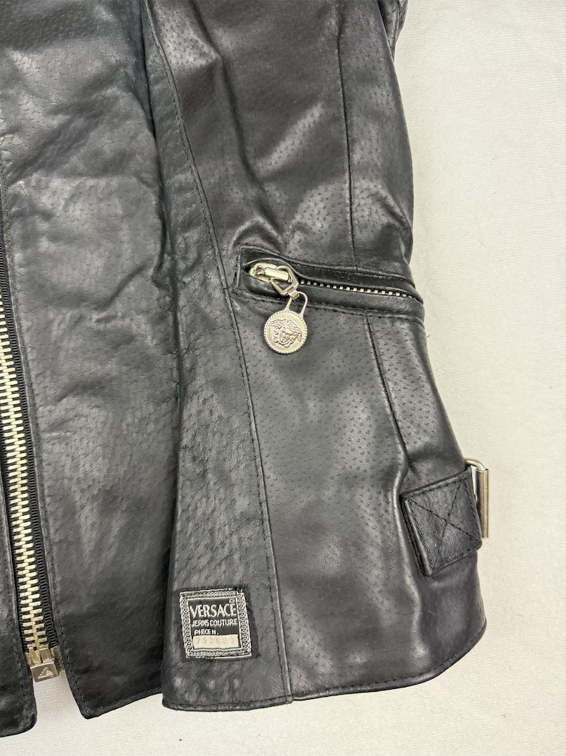 May include: Black leather jacket with a perforated design. Features a silver zipper, a small zippered pocket, and a decorative silver charm. The jacket has a rectangular label with the Versace Jeans Couture logo.