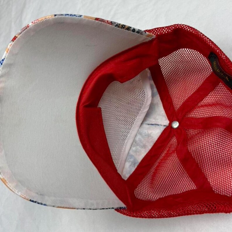 May include: A red and white trucker hat with a mesh back and a patterned front panel. The interior features a white sweatband and a red satin lining. The hat has a snap closure and a curved brim.