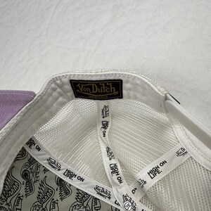 May include: White mesh-backed cap with a purple brim. The interior features a repeating eye design and the text "HIGH ON LIFE." The cap has a black and gold "Von Dutch Originals" label.