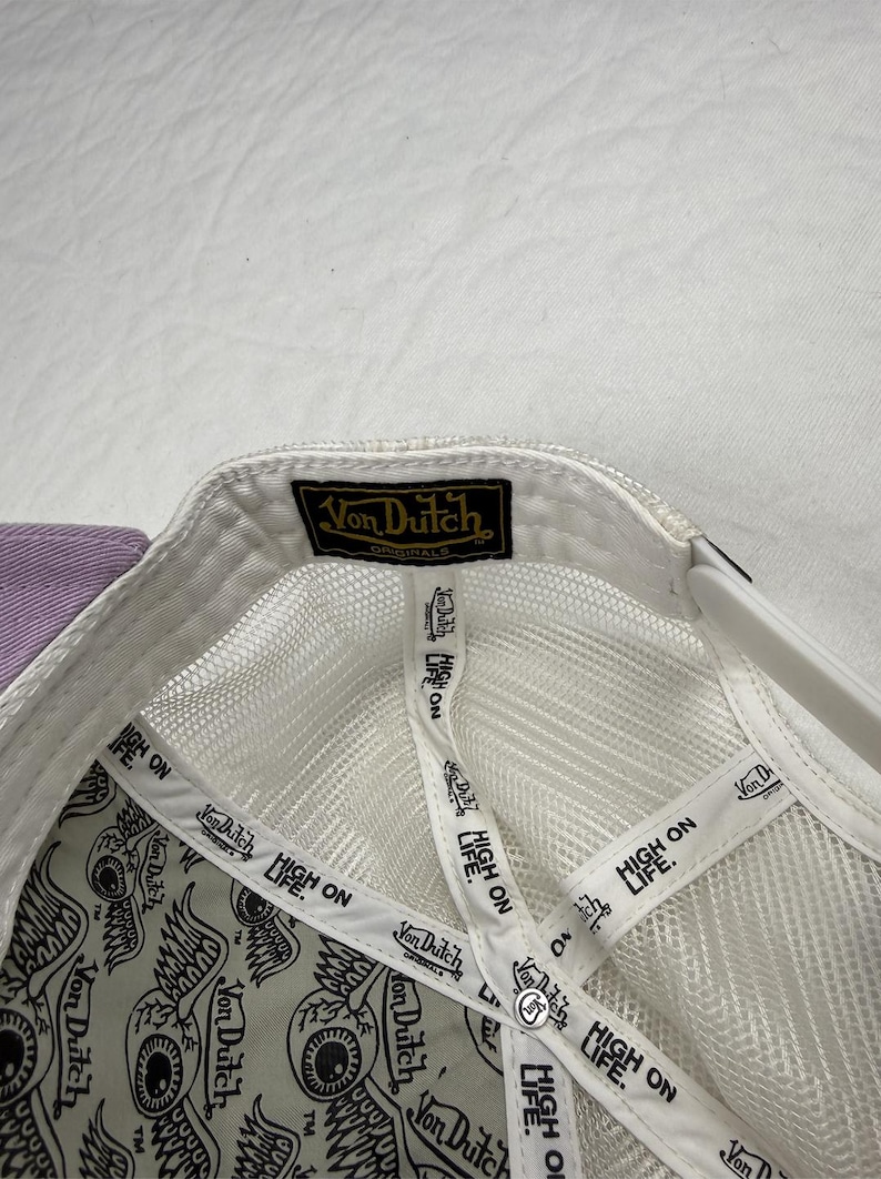 May include: White mesh-backed cap with a purple brim. The interior features a repeating eye design and the text "HIGH ON LIFE." The cap has a black and gold "Von Dutch Originals" label.
