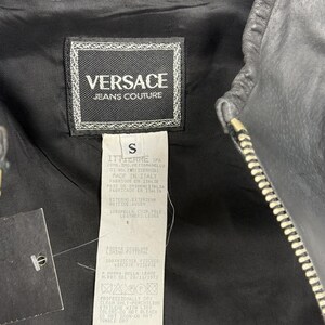 May include: Close-up of a black leather jacket with a silver zipper. The jacket's interior lining is black, and a tag with the "Versace Jeans Couture" logo is visible. The tag also includes size and care instructions.