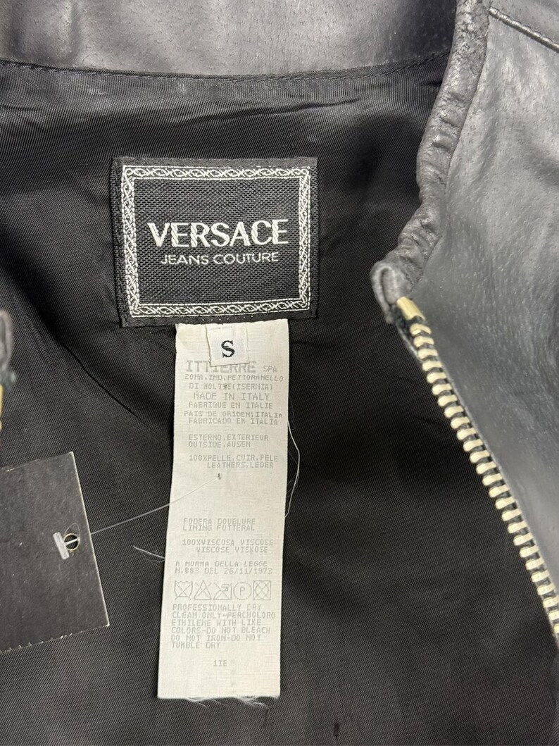 May include: Close-up of a black leather jacket with a silver zipper. The jacket's interior lining is black, and a tag with the "Versace Jeans Couture" logo is visible. The tag also includes size and care instructions.