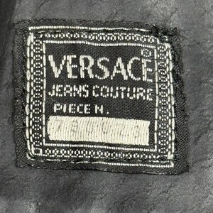 May include: A black and white Versace Jeans Couture label. The label is rectangular with the brand name in large white capital letters. The words "JEANS COUTURE" and "PIECE N." are also visible. The label is stitched onto a black fabric.
