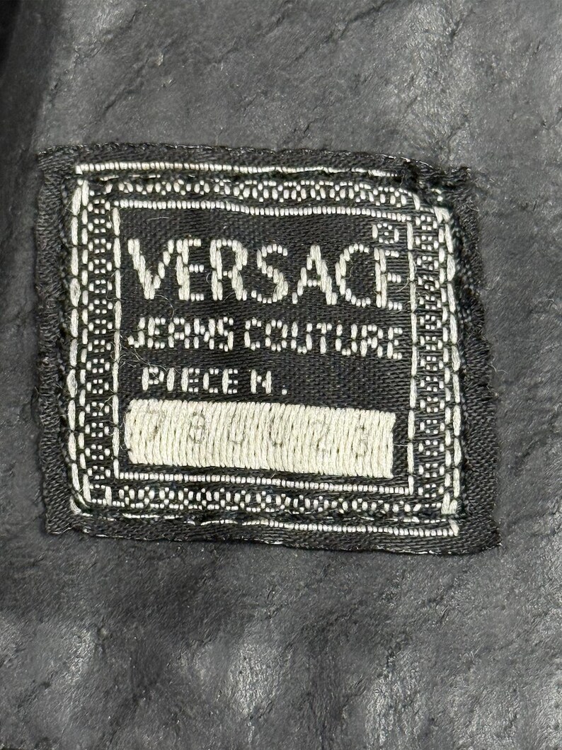 May include: A black and white Versace Jeans Couture label. The label is rectangular with the brand name in large white capital letters. The words "JEANS COUTURE" and "PIECE N." are also visible. The label is stitched onto a black fabric.