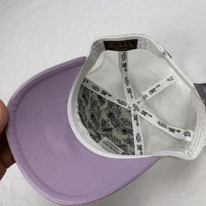 May include: A lavender baseball cap with a white mesh back and a curved brim. The interior features a patterned lining and white straps. The Von Dutch logo is visible on the label.