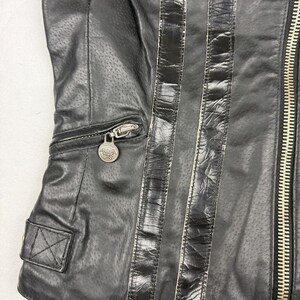 May include: Black leather jacket with perforated detailing and vertical black stripes. Features a silver zipper, a small pocket with a decorative silver medallion, and a small square pocket with a button.