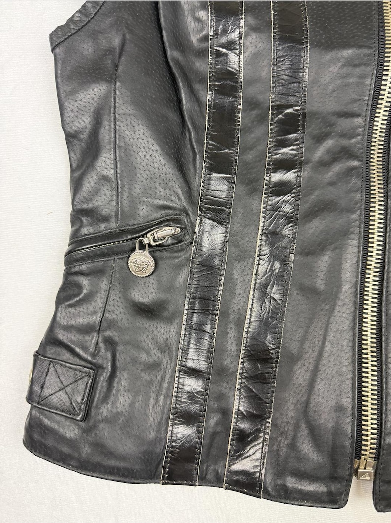 May include: Black leather jacket with perforated detailing and vertical black stripes. Features a silver zipper, a small pocket with a decorative silver medallion, and a small square pocket with a button.