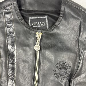 May include: Black leather jacket with a round collar and a silver zipper. The jacket features the Versace Jeans Couture logo on the label and a circular emblem on the chest. The jacket has a perforated design.