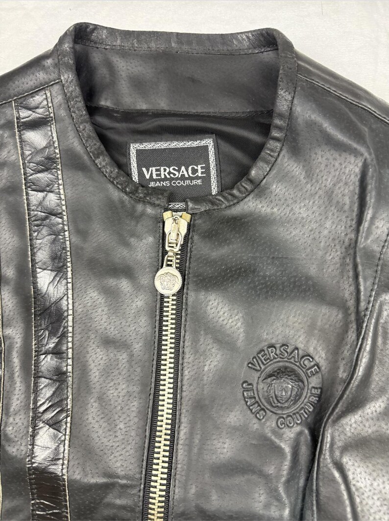 May include: Black leather jacket with a round collar and a silver zipper. The jacket features the Versace Jeans Couture logo on the label and a circular emblem on the chest. The jacket has a perforated design.