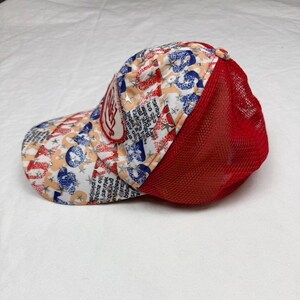 May include: A baseball cap with a red mesh back and a patterned front. The pattern includes red, white, and blue elements with text and star shapes. A patch with text is on the front.