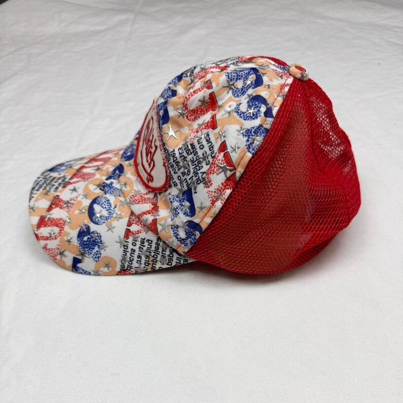 May include: A baseball cap with a red mesh back and a patterned front. The pattern includes red, white, and blue elements with text and star shapes. A patch with text is on the front.