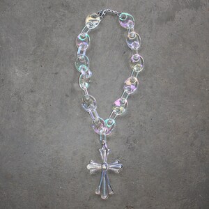 May include: A silver cross pendant hangs from a iridescent clear plastic chain necklace. The necklace is made of large, oval-shaped links.