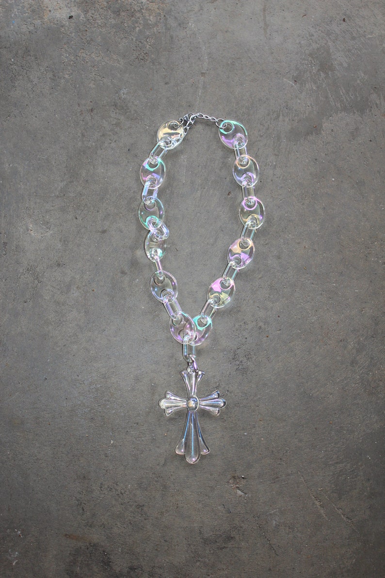 May include: A silver cross pendant hangs from a iridescent clear plastic chain necklace. The necklace is made of large, oval-shaped links.