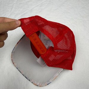 May include: A red mesh trucker hat with an orange adjustable snap closure. The underside of the hat features a white brim with a colorful pattern. The hat is being held up by a hand.