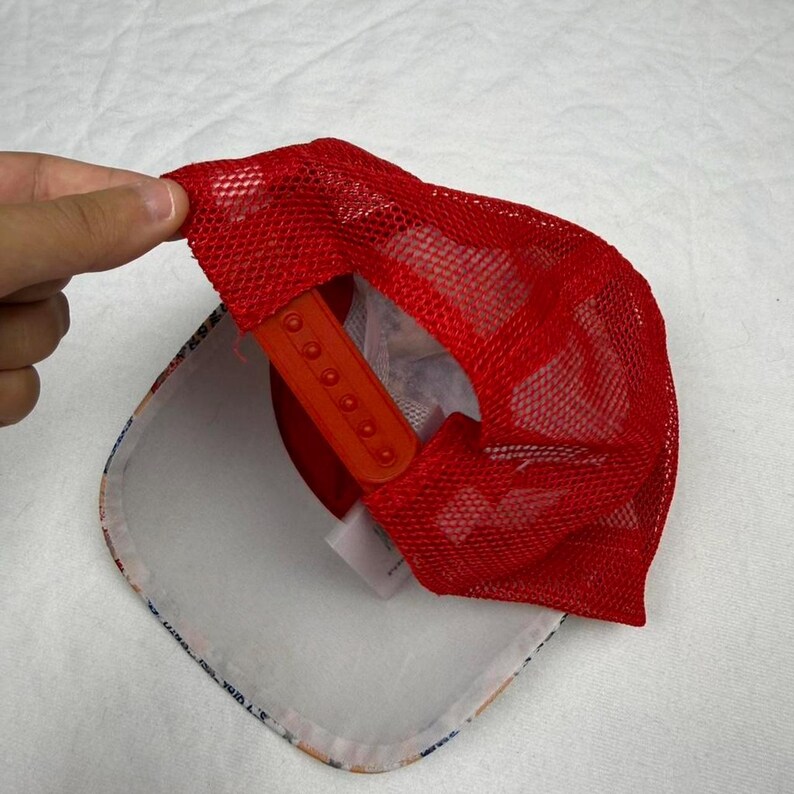 May include: A red mesh trucker hat with an orange adjustable snap closure. The underside of the hat features a white brim with a colorful pattern. The hat is being held up by a hand.