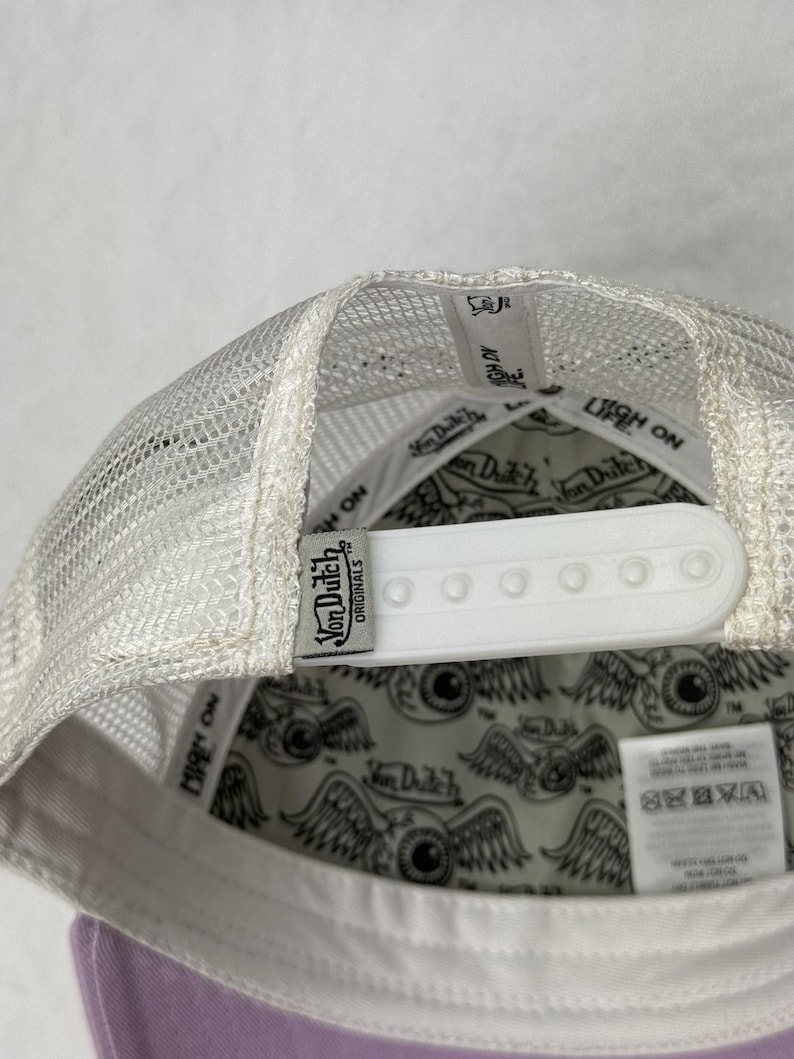 May include: A white mesh-backed baseball cap with a lavender brim. The cap has a white snap closure and a Von Dutch Originals label. The interior lining features a pattern of winged eyeballs.
