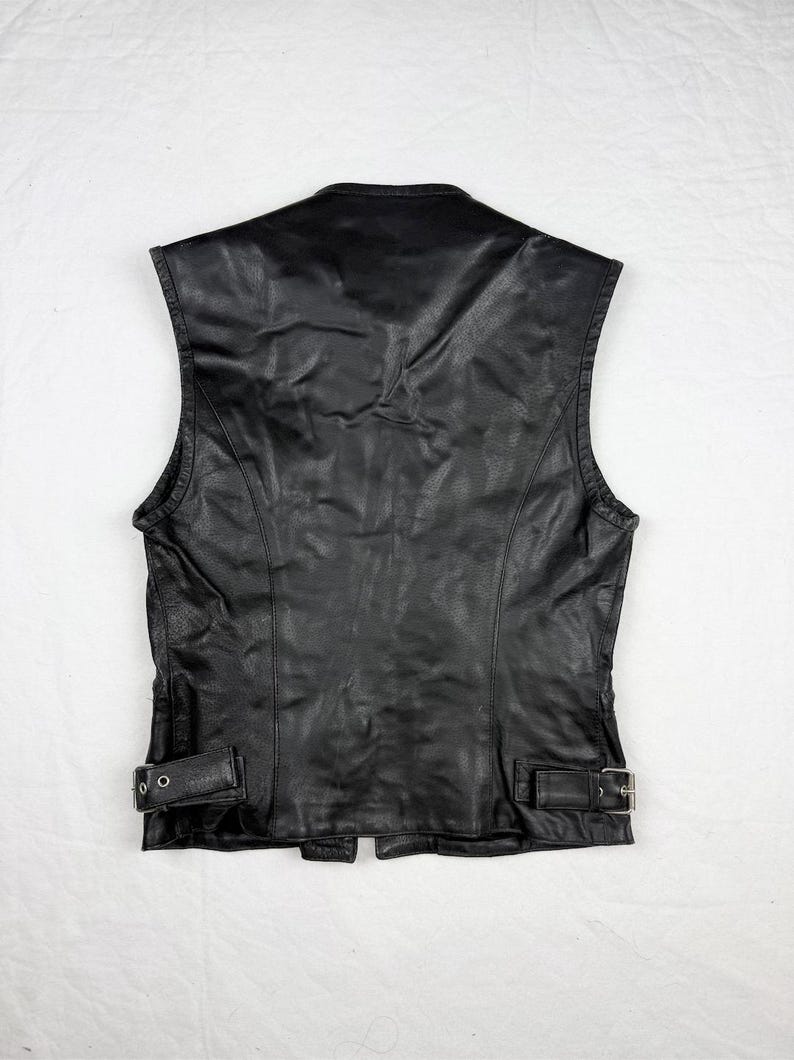 May include: Black leather vest with a classic design. Features a V-neck, sleeveless cut, and adjustable buckles at the waist. The vest has a smooth, slightly textured surface and a tailored fit. A timeless piece for a variety of styles.