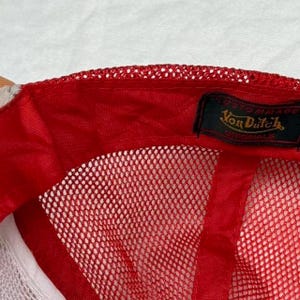 May include: Red and white trucker hat with a mesh back and a curved brim. The hat features a red front panel and a white mesh back. The Von Dutch logo is visible on a black label.